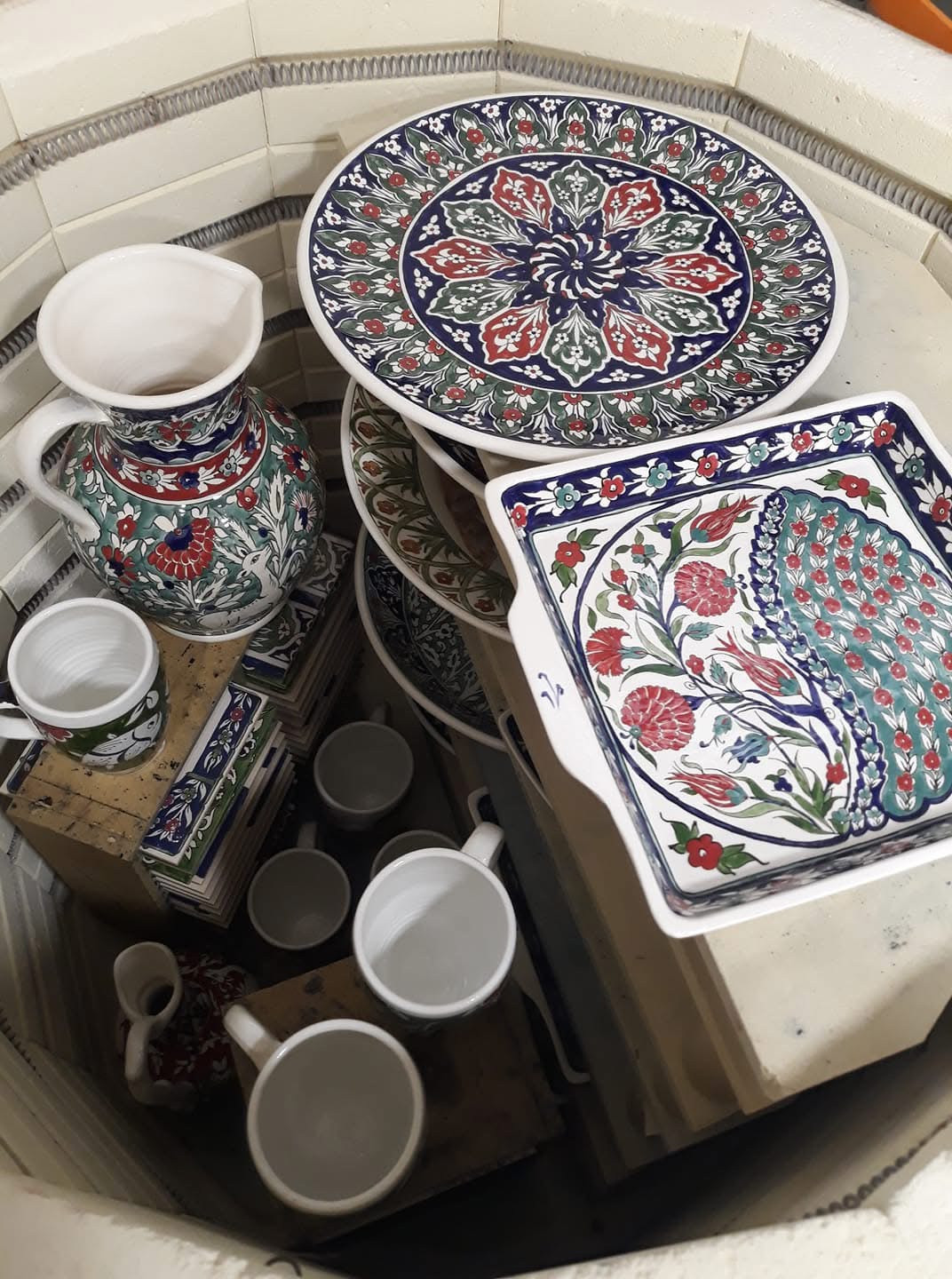 Decorated ceramic plates, a vase and mugs arranged on shelves inside the workshop kiln, with heating coils visible along its walls